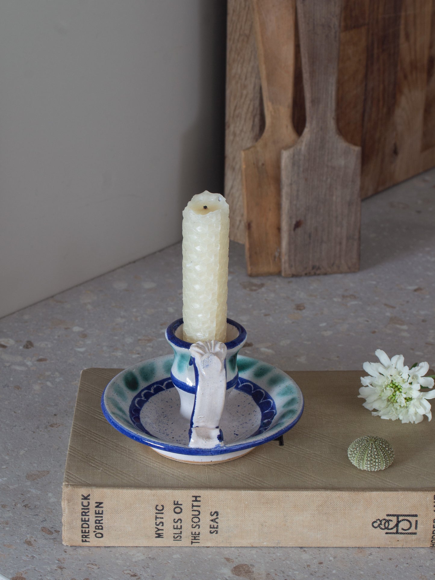 A handmade ceramic candlestick holder with a blue and white patterned saucer and a white pillar candle, styled on a vintage book next to a small white flower.