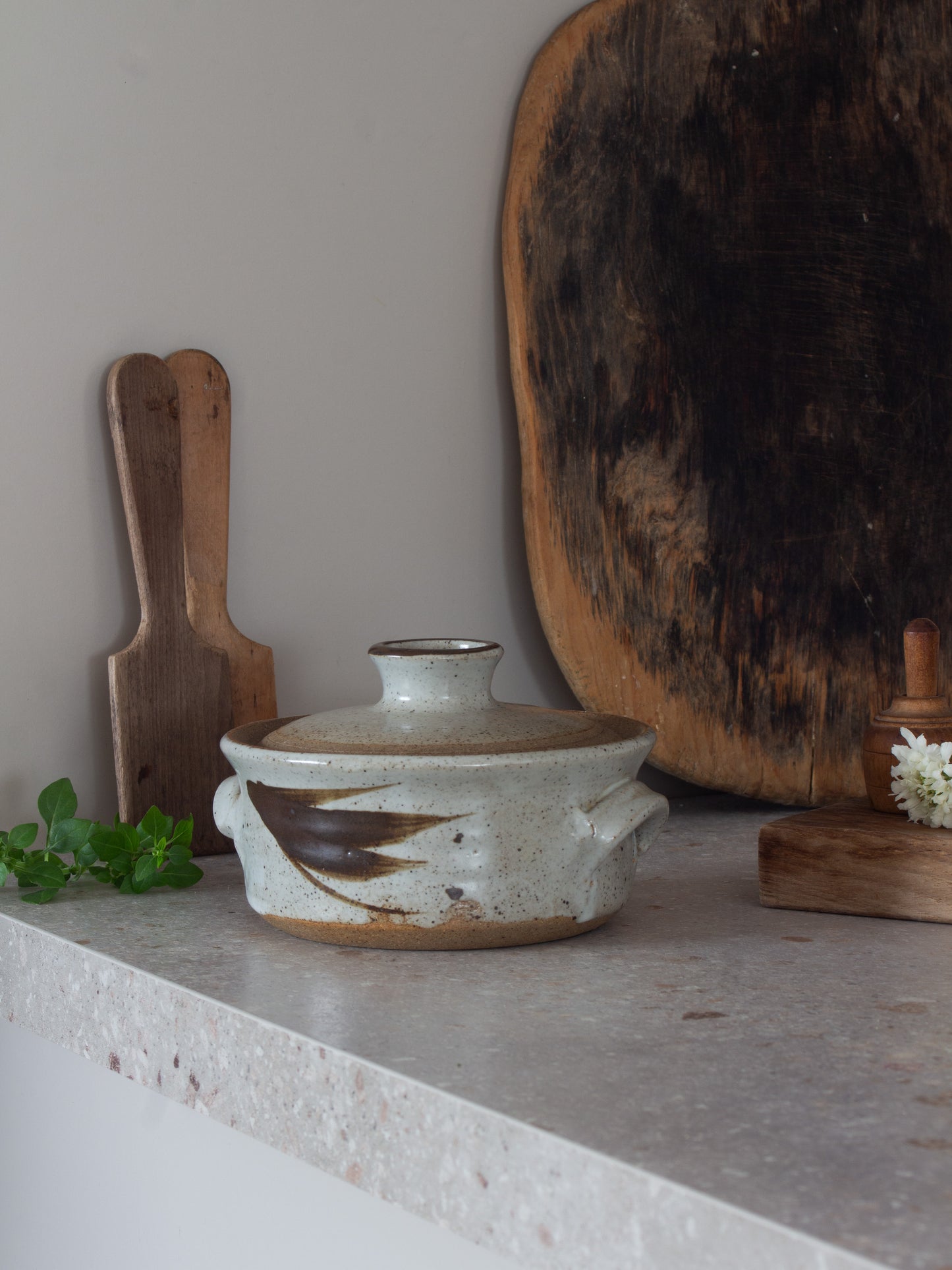 A handmade ceramic lidded casserole dish with a speckled off-white glaze and a dark brown painted accent, styled on a stone countertop with wooden cutting boards and greenery.