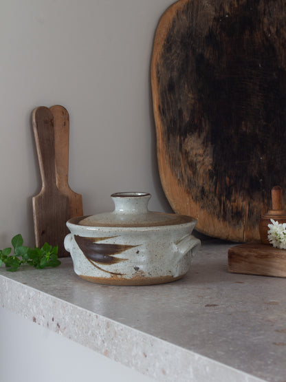 A handmade ceramic lidded casserole dish with a speckled off-white glaze and a dark brown painted accent, styled on a stone countertop with wooden cutting boards and greenery.