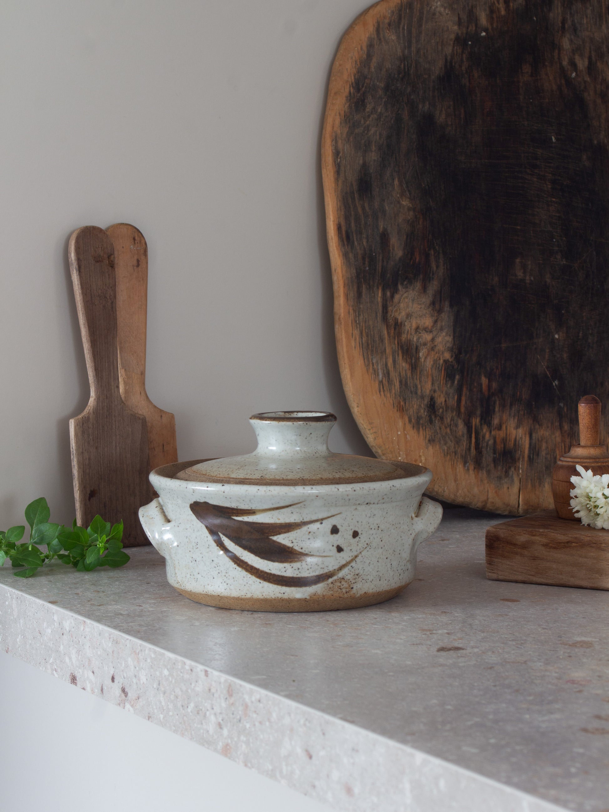 A handmade ceramic lidded casserole dish with side handles and a fish motif, displayed on a stone countertop with rustic wooden cutting boards and herbs in the background.