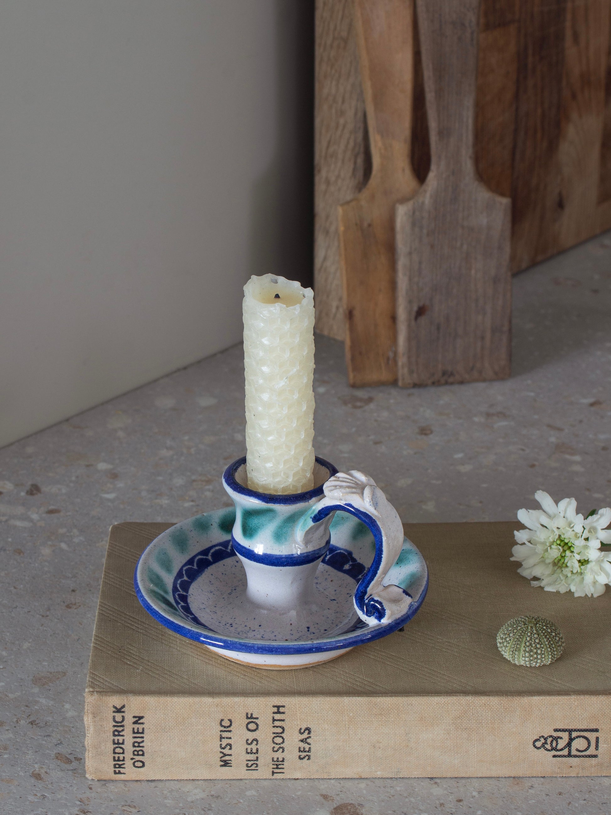 A ceramic candle holder with a sculptural handle and matching saucer, featuring a blue and white glaze with a textured cream candle inserted. The holder is styled on a vintage book with a small white flower and a seashell nearby, against a neutral background.