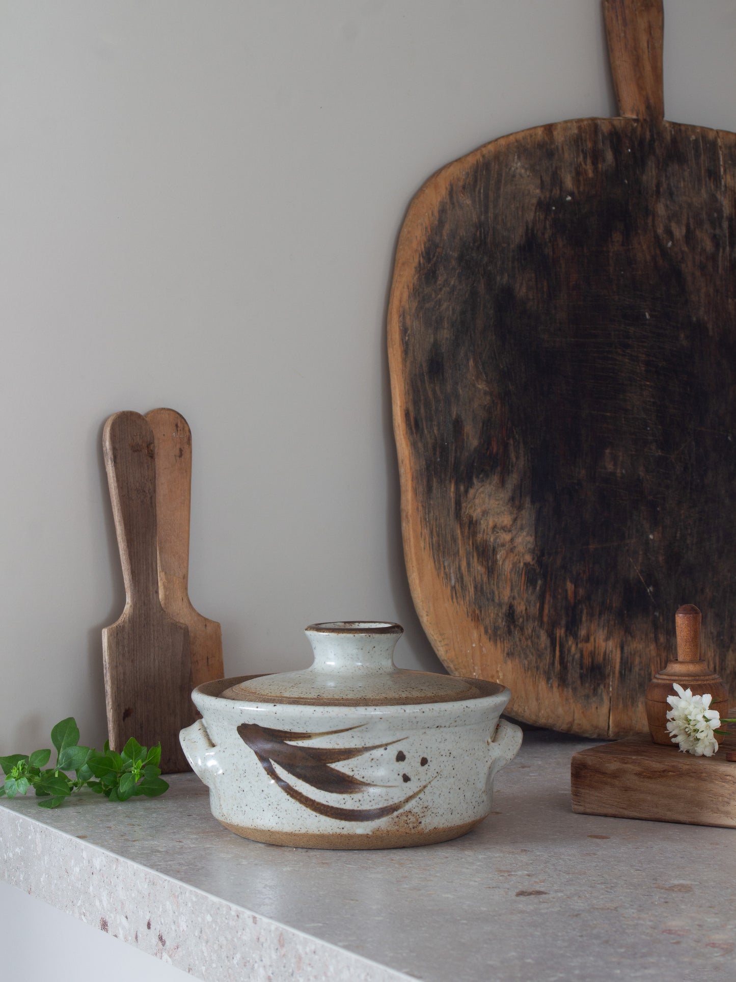 A handmade ceramic lidded casserole dish with side handles and a brown abstract leaf design, displayed on a stone countertop next to rustic wooden cutting boards and a small white flower.