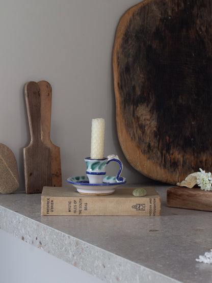 A small white candle in a blue and green patterned ceramic cup and saucer holder, styled on a vintage book on a stone shelf with rustic wooden props.