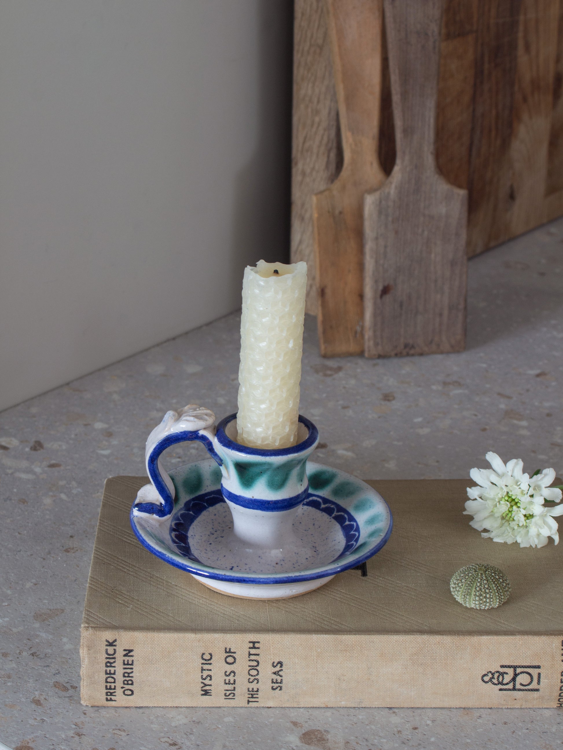 A ceramic candle holder with a handle and saucer, holding a white candle, styled on a vintage book with a small white flower and a seashell against a neutral background.